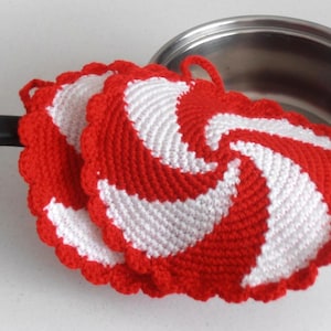 Może przedstawiać: Dwa czerwono-białe szydełkowe podkładki pod garnki ze spiralnym wzorem, obok garnka ze stali nierdzewnej. Podkładki mają falisty brzeg i pętlę do zawieszania. Dekoracja kuchni i domu.