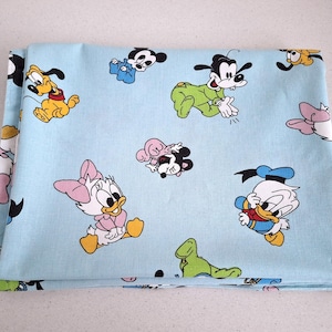 Pode incluir: Tecido azul claro com personagens de desenhos animados, incluindo Mickey Mouse, Minnie Mouse, Pato Donald, Margarida, Pateta e Pluto. O tecido está dobrado. Os personagens estão em várias poses.