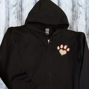 May include: Black zippered hoodie with a red and white paw print with a heart in the center. The hoodie has a hood and long sleeves. The text "Pet Themed Shirts Made in the USA Mydeye" is on a wooden sign below the hoodie.