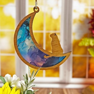 May include: A wooden crescent moon ornament with a blue, purple, and pink abstract design. A wooden cat silhouette sits on the moon.