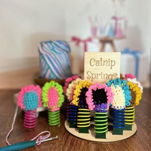 May include: A wooden stand with a sign that reads "Catnip Springs" holds colorful crocheted flowers attached to metal springs.
