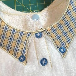 Puede incluir: Primer plano de un babero de tela blanca y afelpada con un cuello a cuadros en amarillo, azul y crema. El cuello está ribeteado con un hilo dorado y tiene tres botones azules con detalles bordados.