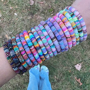 Puede incluir: Una pila de pulseras de cuentas coloridas en varios tonos de morado, azul, rosa y verde. Las pulseras están hechas de cuentas pequeñas, estrechamente tejidas y están dispuestas en un orden de tamaño graduado, con la pulsera más grande en la parte inferior y la más pequeña en la parte superior.