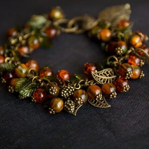 May include: A handcrafted bracelet with amber-coloured beads, green leaf accents, and bronze-toned metalwork. The design incorporates small, textured beads and decorative leaf charms, creating a rustic, autumnal aesthetic. The bracelet is a unique piece of jewellery.