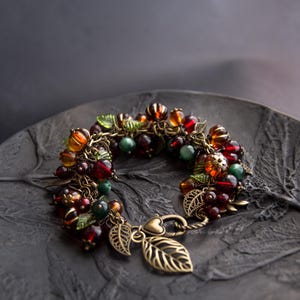May include: A handcrafted bracelet with a cluster of red, orange, and green beads. The bracelet has leaf and heart-shaped charms, and is made with antique bronze-toned metal. The bracelet is displayed on a dark grey leaf-shaped plate.
