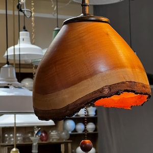 Puede incluir: Una lámpara colgante de madera con un acabado de madera natural y un cálido brillo naranja. La luz cuelga de un cable negro y tiene un interruptor de cadena.