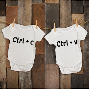 Puede incluir: Dos bodies blancos para bebés colgados de una cuerda para tender la ropa. El body de la izquierda dice "Ctrl + c" y el body de la derecha dice "Ctrl + v".