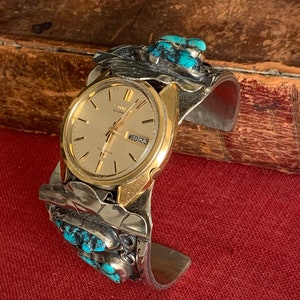 May include: A gold-toned wristwatch with a silver cuff bracelet. The cuff is decorated with turquoise stones and silver accents. The watch face displays the date and day of the week. The watch is on a red surface.
