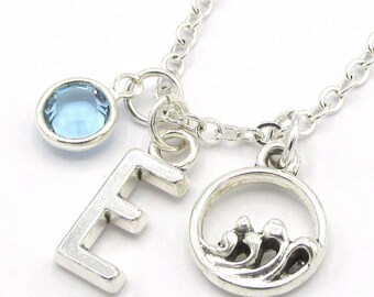 May include: A silver chain necklace with a blue crystal charm, a silver letter E charm, and a silver charm of a wave in a circle.