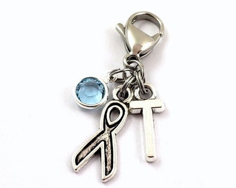 May include: A silver charm featuring a blue crystal, a silver ribbon, and a silver letter T. The charm is attached to a silver lobster clasp.