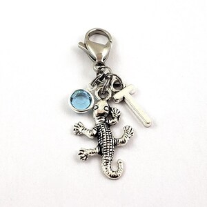 May include: A silver charm with a blue crystal, a silver lizard charm, and a silver letter T charm. The charms are attached to a silver lobster clasp.