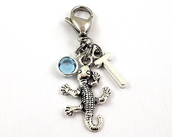 May include: A silver charm with a blue crystal, a silver lizard charm, and a silver letter T charm. The charms are attached to a silver lobster clasp.