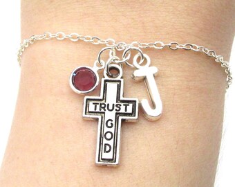 May include: Silver charm bracelet featuring a cross charm inscribed with "Trust God", a letter J charm, and a small red gemstone charm.