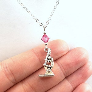 May include: A silver microscope charm necklace with a pink crystal accent. The necklace is on a silver chain.
