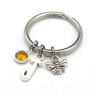May include: A silver key chain with a bee charm, a letter J charm, and a small amber-colored rhinestone.