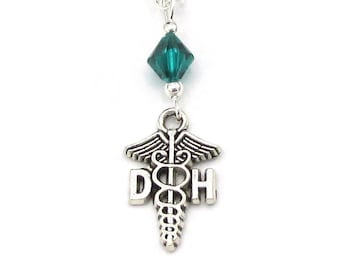 May include: A silver-toned necklace featuring a pendant with the letters "DOH" and a medical symbol, suspended from a delicate chain. A faceted teal crystal bead accents the design. The necklace is displayed against a white background.
