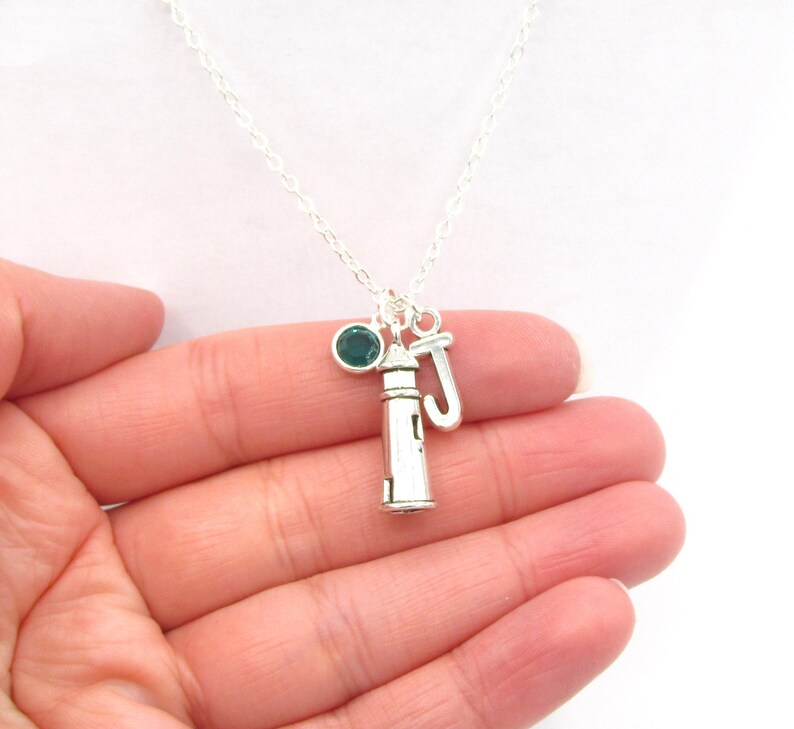 May include: Silver necklace featuring a lighthouse charm, a small green gemstone, and a letter 'J' charm.