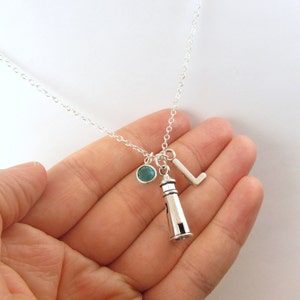 May include: Silver necklace with a lighthouse charm, a letter L charm, and a teal blue crystal charm.