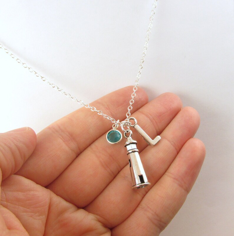 May include: Silver necklace with a lighthouse charm, a letter L charm, and a teal blue crystal charm.
