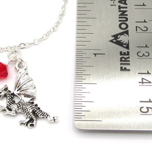 May include: Silver dragon charm necklace with a red crystal bead. The charm is about 1.5 inches tall and is hanging from a silver chain. The necklace is next to a ruler for size reference.