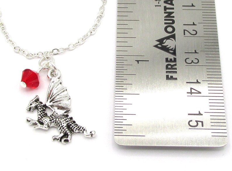 May include: Silver dragon charm necklace with a red crystal bead. The charm is about 1.5 inches tall and is hanging from a silver chain. The necklace is next to a ruler for size reference.