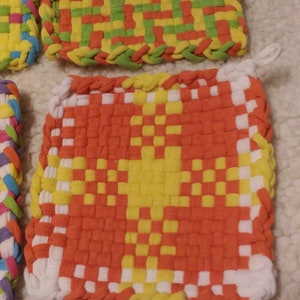 May include: Four colorful woven pot holders with a variety of patterns. The pot holders are made of fabric strips woven together in a checkered pattern. The colors include red, orange, yellow, green, blue, and white.