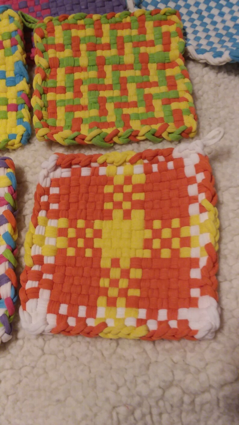 May include: Four colorful woven pot holders with a variety of patterns. The pot holders are made of fabric strips woven together in a checkered pattern. The colors include red, orange, yellow, green, blue, and white.