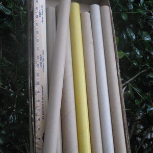 May include: A cardboard box filled with several cylindrical tubes of varying colors, including beige, yellow, and white. A wooden ruler with printed text is placed inside the box, next to the tubes. The tubes appear to be for art or craft projects.