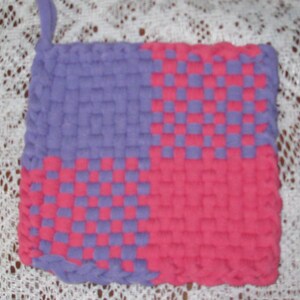 May include: A purple and pink woven pot holder with a checkered pattern. The pot holder is made of a soft, textured fabric.