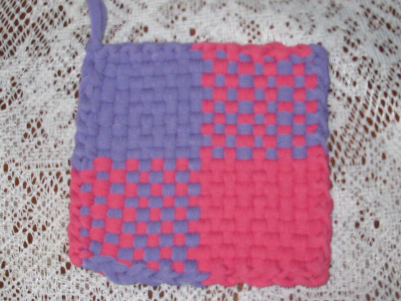 May include: A purple and pink woven pot holder with a checkered pattern. The pot holder is made of a soft, textured fabric.