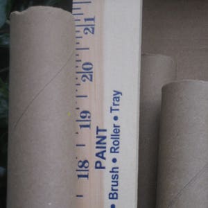 May include: A close-up shot features a wooden ruler with inch markings and the text "PAINT Brush Roller Tray". Two cardboard tubes are visible, one next to the ruler and two others in the background. The ruler measures up to 22 inches.