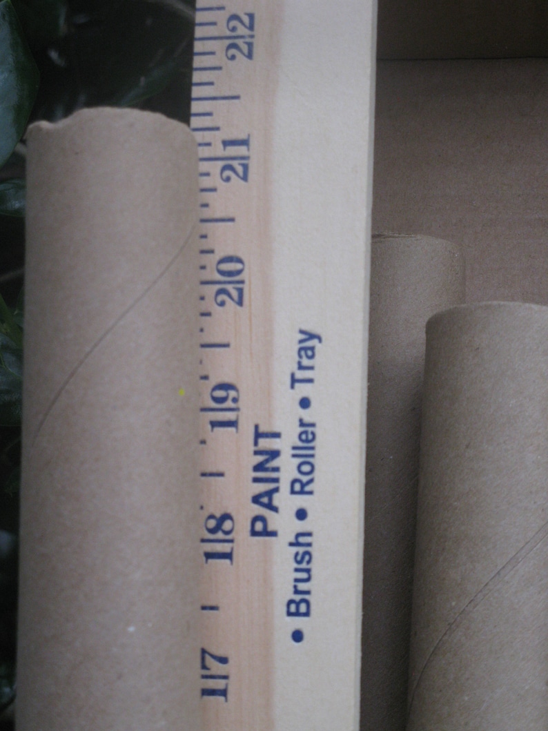 May include: A close-up shot features a wooden ruler with inch markings and the text "PAINT Brush Roller Tray". Two cardboard tubes are visible, one next to the ruler and two others in the background. The ruler measures up to 22 inches.