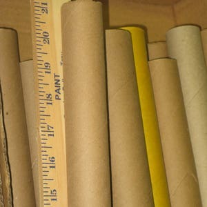 May include: A collection of cardboard tubes of varying sizes and colors, including brown and yellow. A wooden ruler with inch measurements is visible, with the numbers 14 through 22. The tubes are inside a cardboard box.