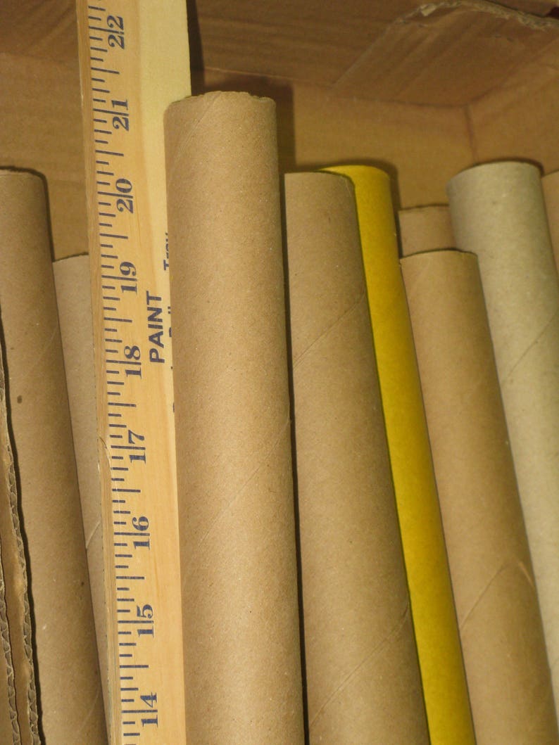 May include: A collection of cardboard tubes of varying sizes and colors, including brown and yellow. A wooden ruler with inch measurements is visible, with the numbers 14 through 22. The tubes are inside a cardboard box.