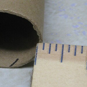 May include: A close-up of a cardboard tube with a ruler. The tube is brown and has a dark interior. The ruler is wooden with blue markings, showing a measurement of approximately 1 inch.