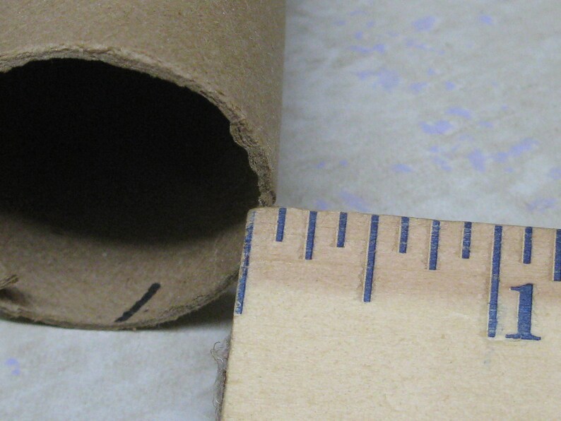 May include: A close-up of a cardboard tube with a ruler. The tube is brown and has a dark interior. The ruler is wooden with blue markings, showing a measurement of approximately 1 inch.