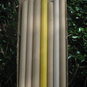 May include: A cardboard box filled with several cylindrical paper tubes. The tubes are various shades of tan and gray, with one bright yellow tube in the center. The tubes are arranged vertically.