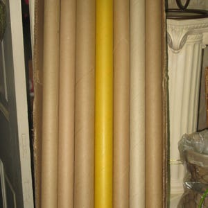 May include: A collection of cylindrical cardboard tubes, varying in shades of brown and tan, are arranged inside a cardboard box. One tube stands out with a bright yellow hue, contrasting with the neutral tones of the others. The tubes are vertically oriented.