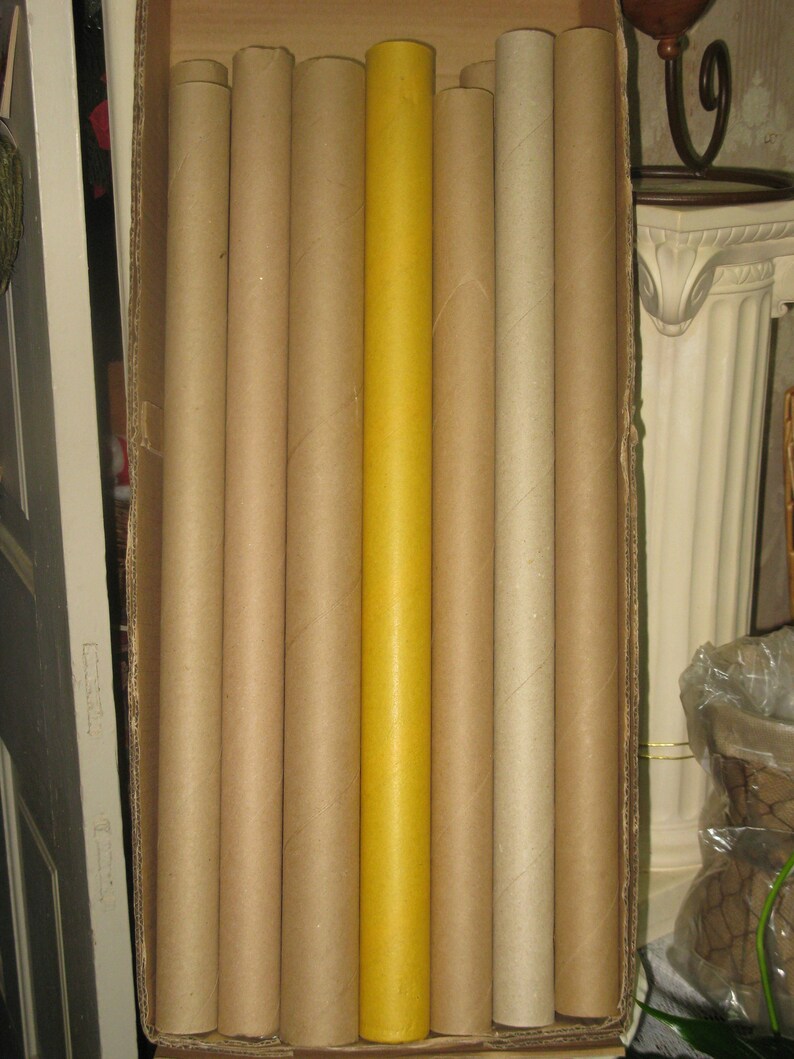 May include: A collection of cylindrical cardboard tubes, varying in shades of brown and tan, are arranged inside a cardboard box. One tube stands out with a bright yellow hue, contrasting with the neutral tones of the others. The tubes are vertically oriented.