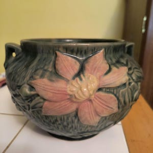 May include: A green ceramic planter with a raised floral design. The planter features a large pink flower with a yellow center and green leaves.