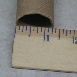May include: A brown cardboard tube with a diameter of approximately 1 inch is positioned next to a wooden ruler. The ruler displays inch markings in blue ink. The tube's end is visible, showing its hollow interior.