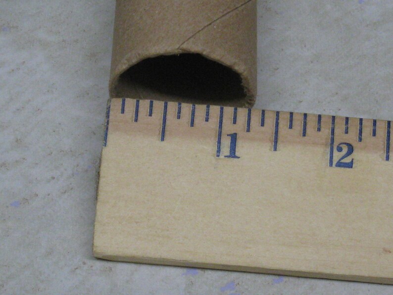 May include: A brown cardboard tube with a diameter of approximately 1 inch is positioned next to a wooden ruler. The ruler displays inch markings in blue ink. The tube's end is visible, showing its hollow interior.