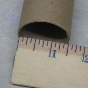 May include: A brown cardboard tube with a ruler measuring its length. The ruler shows the tube is approximately 1 inch in diameter. The tube is open at one end, revealing a dark interior. The ruler has blue markings.