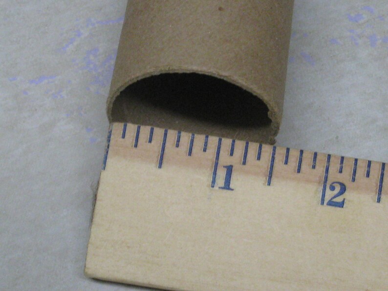 May include: A brown cardboard tube with a ruler measuring its length. The ruler shows the tube is approximately 1 inch in diameter. The tube is open at one end, revealing a dark interior. The ruler has blue markings.