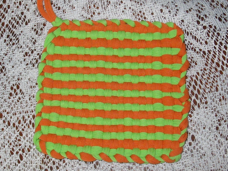 May include: A green and orange woven pot holder made from strips of fabric.