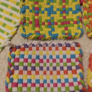 May include: Four colorful woven pot holders with a checkered pattern. The pot holders are made of fabric strips in shades of purple, green, yellow, orange, pink, blue, and white.