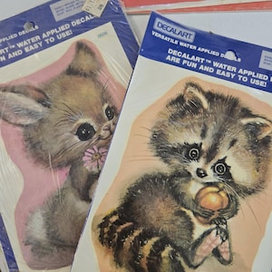 May include: Two packages of Decalart water-applied decals in plastic sleeves. One package features a brown kitten holding a pink flower, while the other displays a raccoon holding a peach. Both packages include blue headers with white text.
