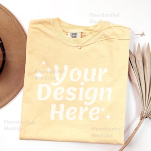 May include: A light yellow t-shirt with the text "Your Design Here" in white, a brown hat, a gold necklace, and a dried palm leaf on a white background. The t-shirt has a Comfort Colors tag.