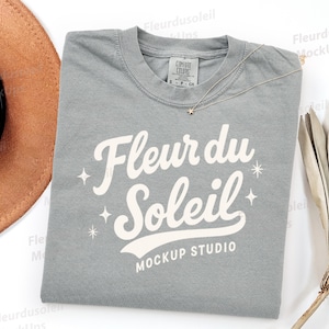 Può includere: Una maglietta grigia con la scritta "Fleur du Soleil" in un carattere bianco e stilizzato. La maglietta ha uno scollo tondo e una piccola etichetta sul colletto. Sono visibili anche una collana d'oro con un ciondolo a stella e un cappello marrone.