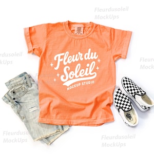 May include: An orange t-shirt with the text "Fleur du Soleil" and a pair of light blue denim shorts are displayed on a white background. A pair of black and white checkered slip-on shoes are also present, creating a casual, summery aesthetic.